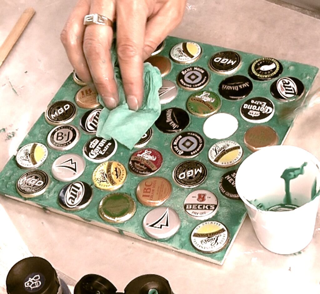 Use paper towel to wipe off excess greenish teal paint from beer bottle cap trivet