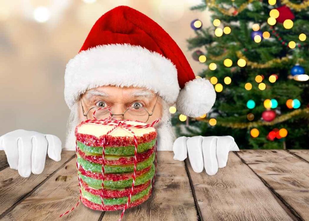 Santa peeking over the table at a stack of christmas cookies with a Christmas tree in the background
