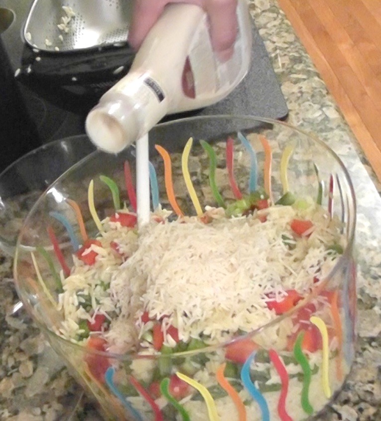 Orzo summer pasta salad ingredients mixed in bowl and pouring dressing on top
