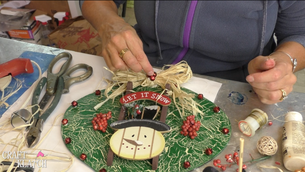 I found this ornament at Dollar Tree, in fact I found four! I decided I would style the ornament four different ways! This is the first and I'm going for a Rustic Snowman Christmas Ornament DIY! Dollar store crafts are a great way to decorate on a budget and customize your Christmas decorations to reflect your personal style and home decor ideas! #craftklatch #holidaydecorations #dollartree #dollartreecrafts #christmas #christmascraft #christmascraftideas #diy #craft #crafts #crafting #craftideas #howto #christmasdecor, Adding the jingle bells to the raffia bow