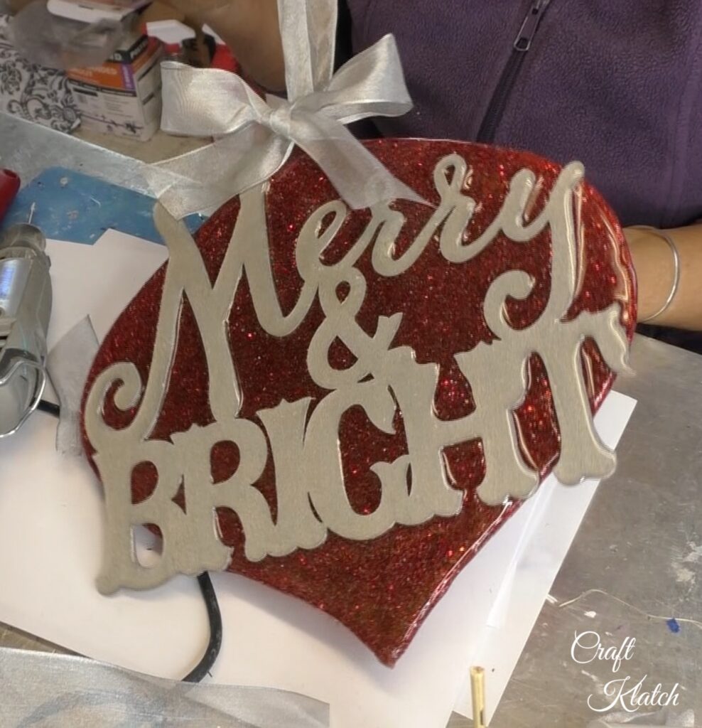 Holding up finished Dollar Tree ornament with red glitter and silver merry and bright and silver bow