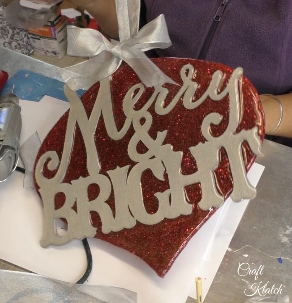 Dollar Tree Ornament Makeover #2 and this time we added glitz and glam to the Christmas decoration tutorial! A little resin and glitter make holiday art! Fun Christmas craft ideas! Dollar store crafts are great for decorating on a budget! #craftklatch #craftideas #chiristmas #christmascrafts #resin #resinart #dollartree #dollartreecrafts #dollartreediys Holding up finished Dollar Tree ornament with red glitter and silver merry and bright and silver bow