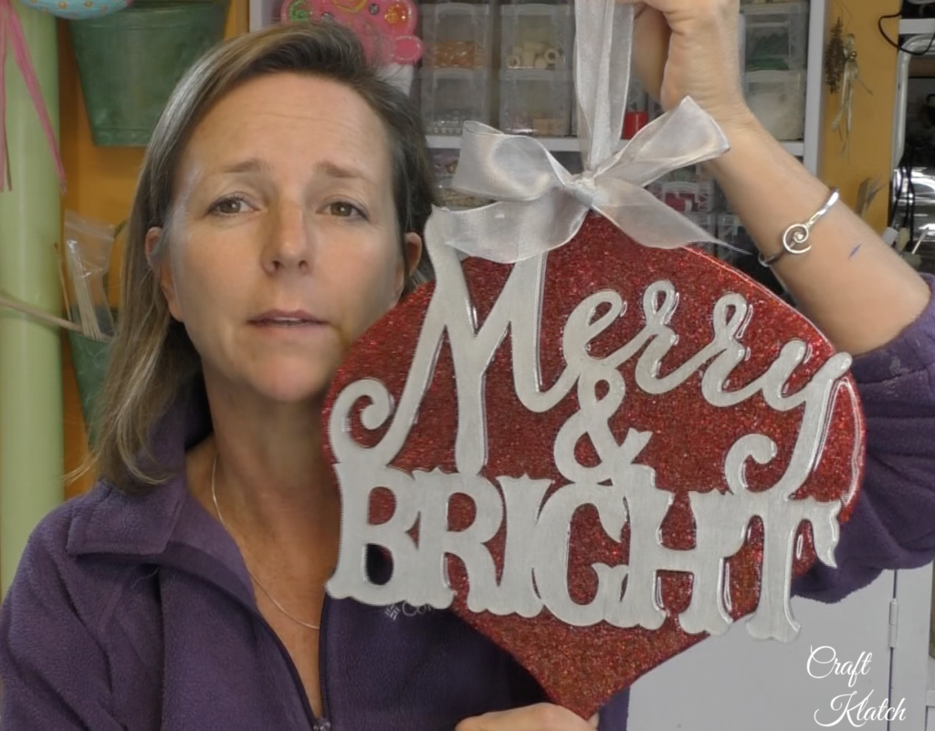 Dollar Tree Ornament Makeover #2 and this time we added glitz and glam to the Christmas decoration tutorial! A little resin and glitter make holiday art! Fun Christmas craft ideas! #craftklatch #craftideas #chiristmas #christmascrafts #resin #resinart #dollartree #dollartreecrafts #dollartreediys Mona of Craft Klatch holding up finished Dollar Tree ornament makeover Merry and Bright