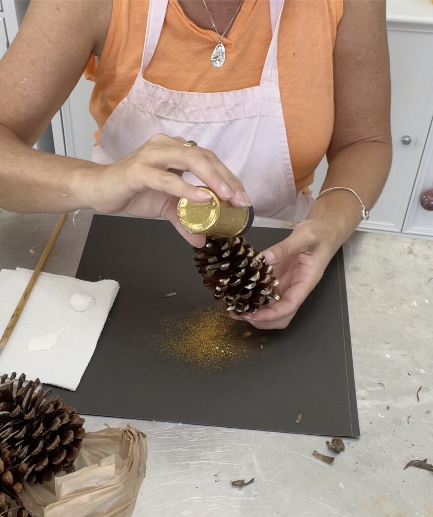 Pine cone craft for Christmas being sprinkled with gold glitter copy